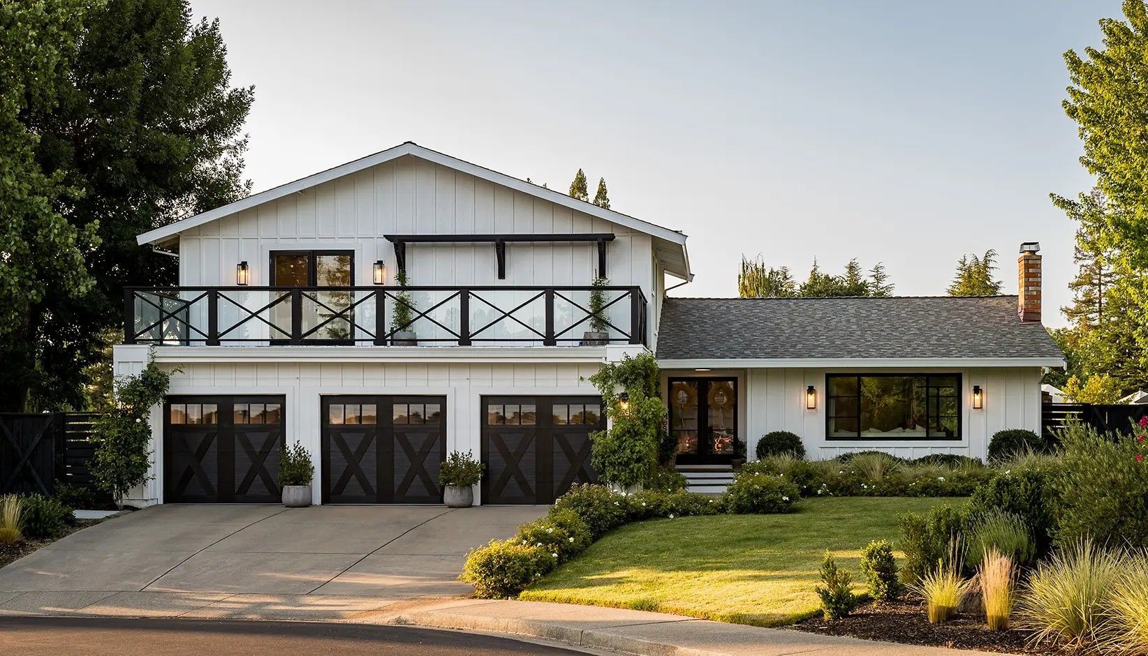 A white two-story suburban house with a large balcony, three dark garage doors, and a lush, manicured front lawn.
