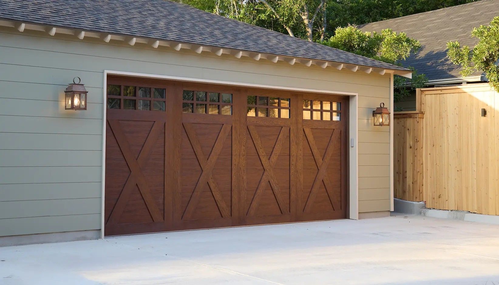 A brown garage door with X-pattern detailing and rectangular windows, installed on a light-colored building with lamps.