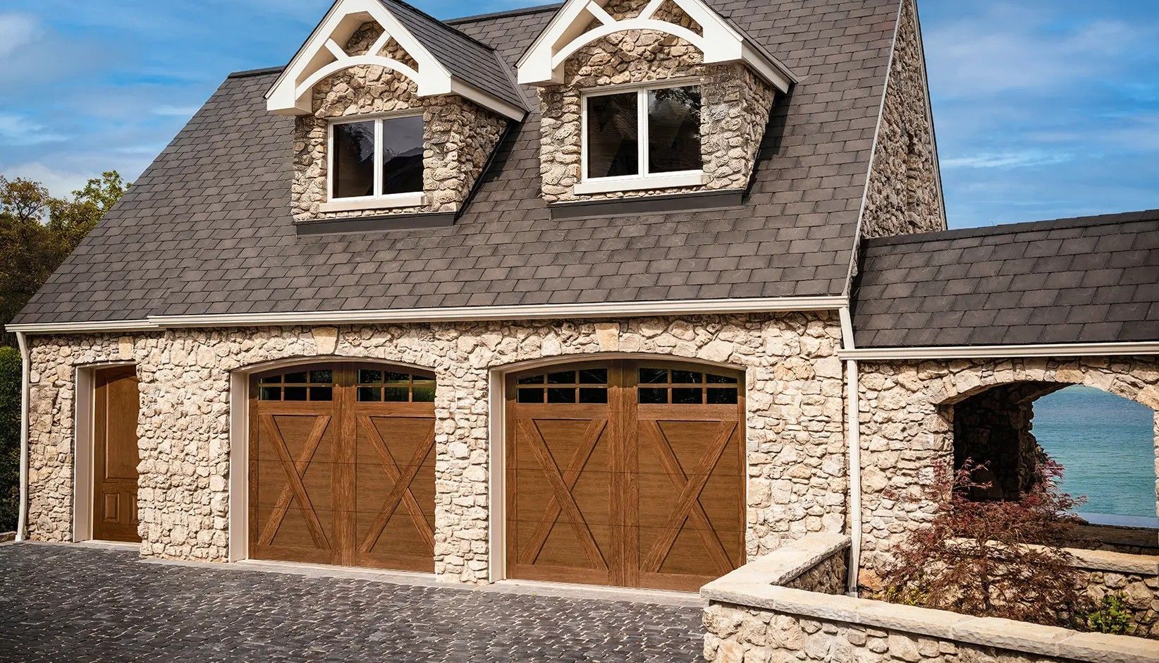 A two-story stone garage with two wooden carriage-style doors and two gabled dormer windows under a dark shingle roof.