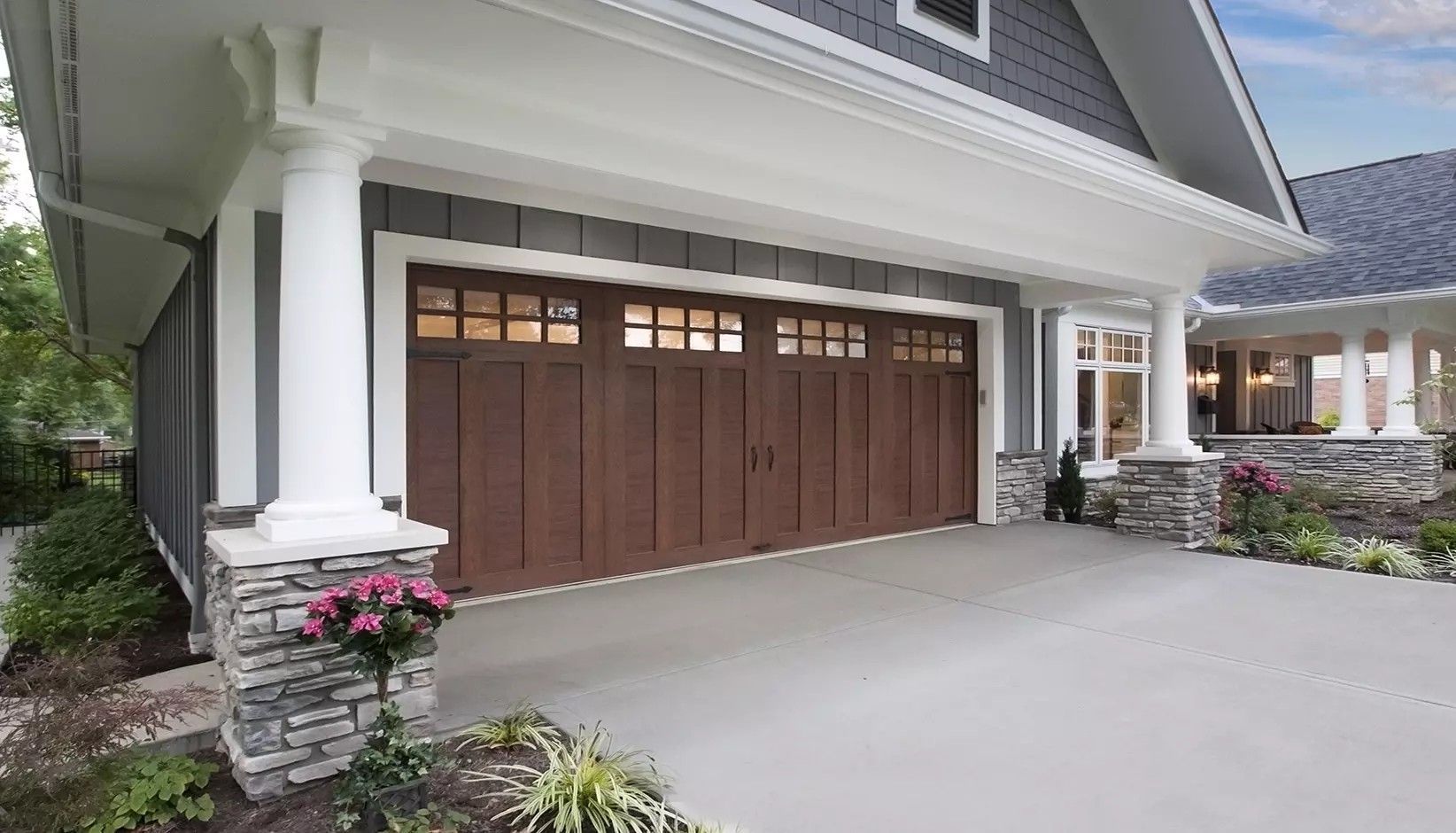 A dark wood garage door with windows on a house with gray siding and stone pillars under a large covered entryway.