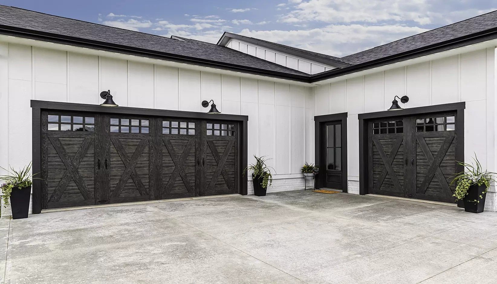 White modern farmhouse exterior with dark wood garage doors, a matching side door, and potted plants on a concrete drive.