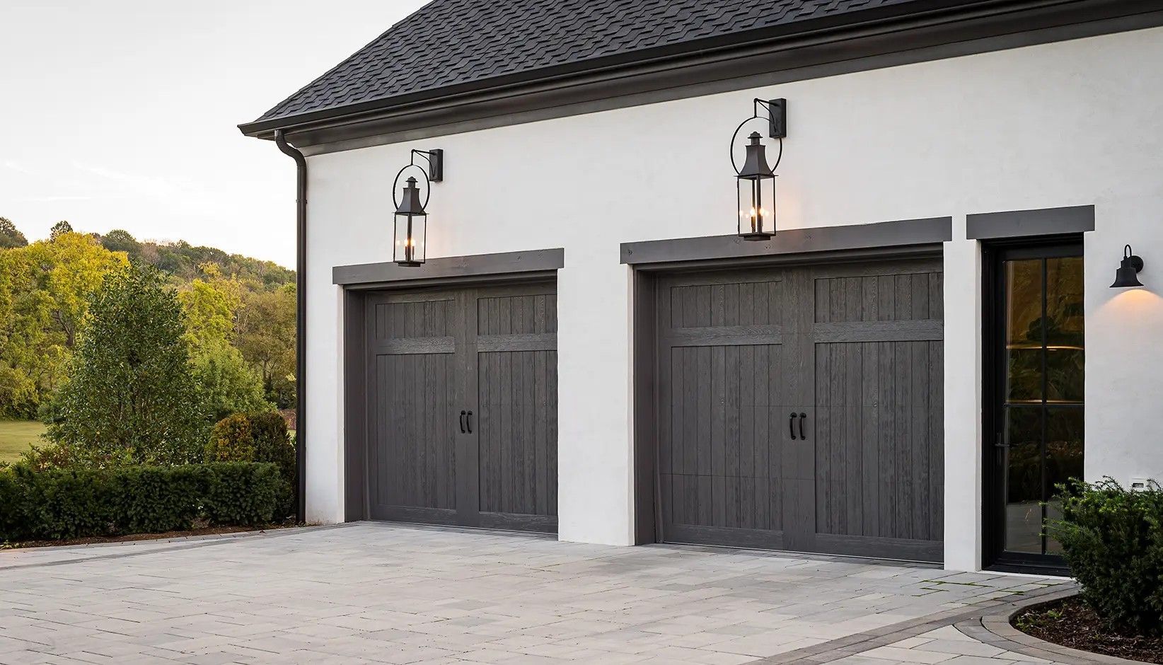 Two dark grey carriage-style garage doors on a white stucco building with outdoor wall lanterns.