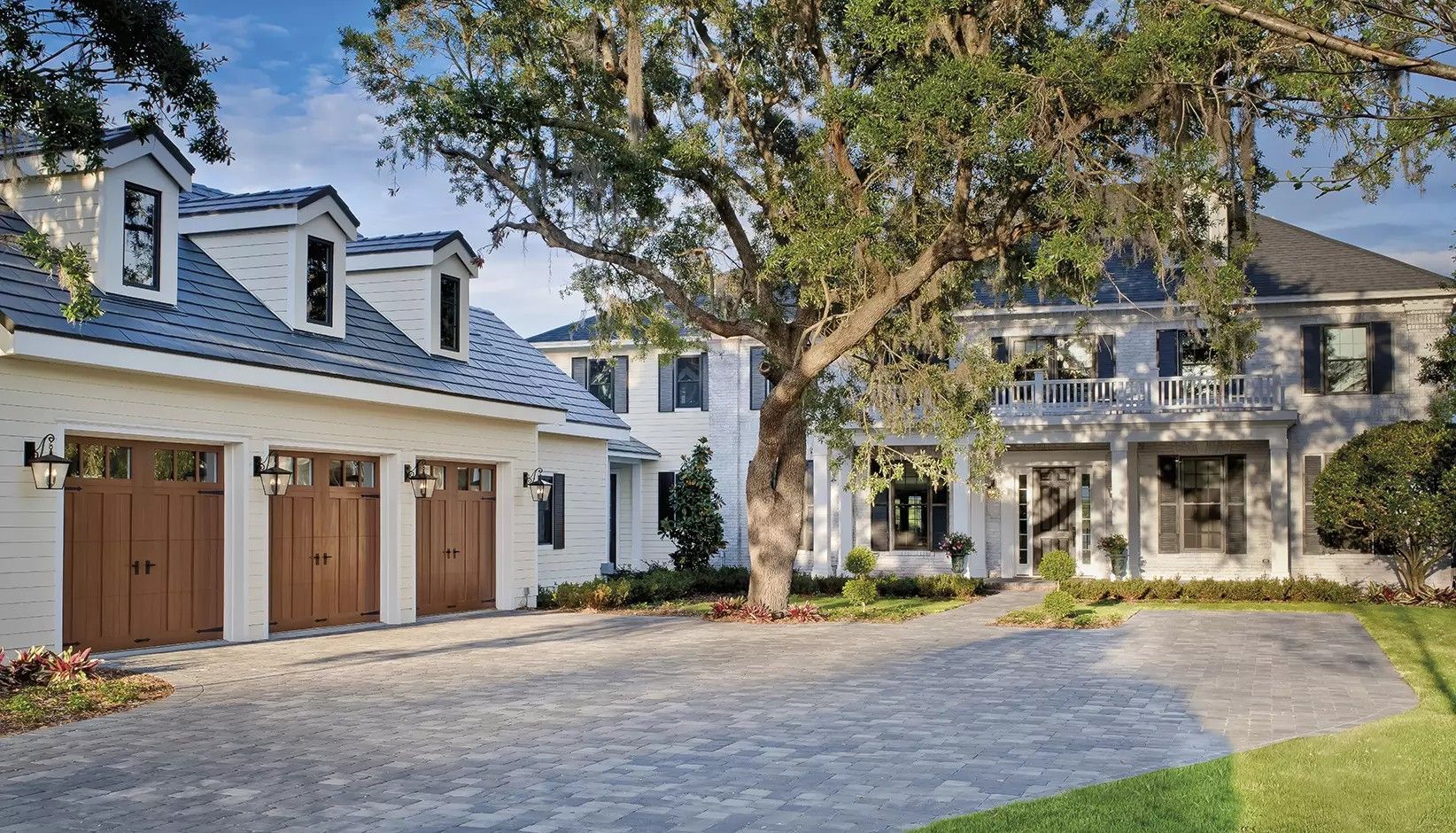 A large white two-story house with a three-car garage and a stone driveway, shaded by a prominent, sprawling oak tree.