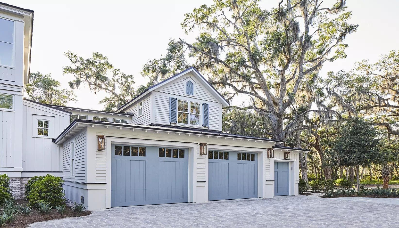 A white house with a multi-bay, light blue garage, wooden trim, and surrounding oak trees on a gravel driveway.