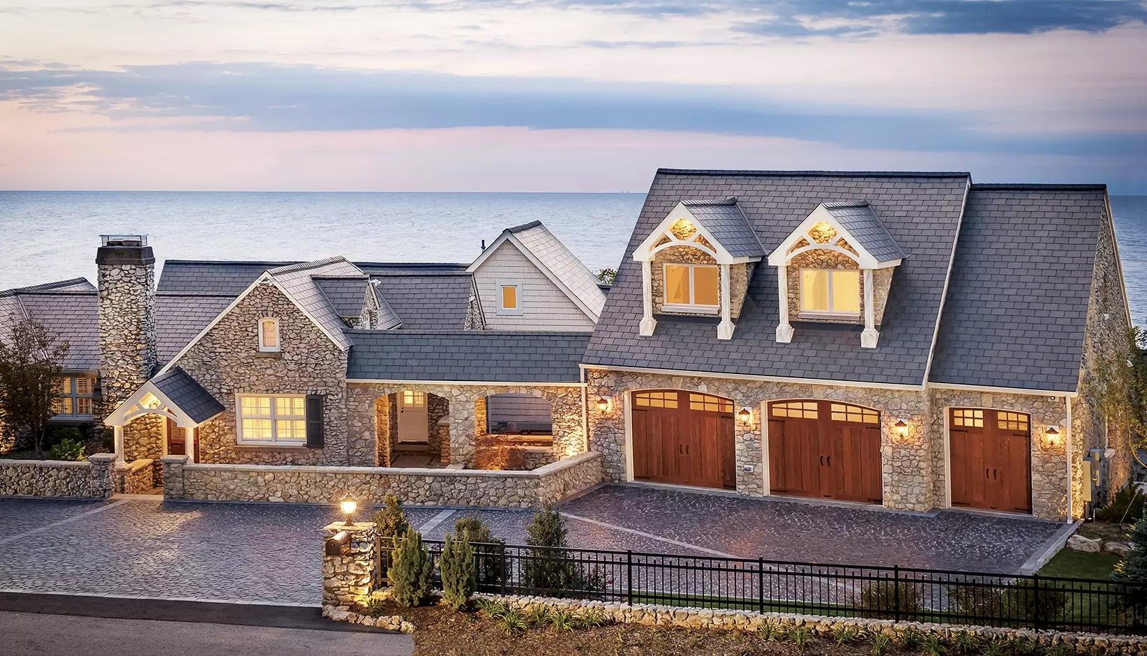 A stone house with three wooden garage doors sits by the ocean at dusk, illuminated by warm outdoor lights.