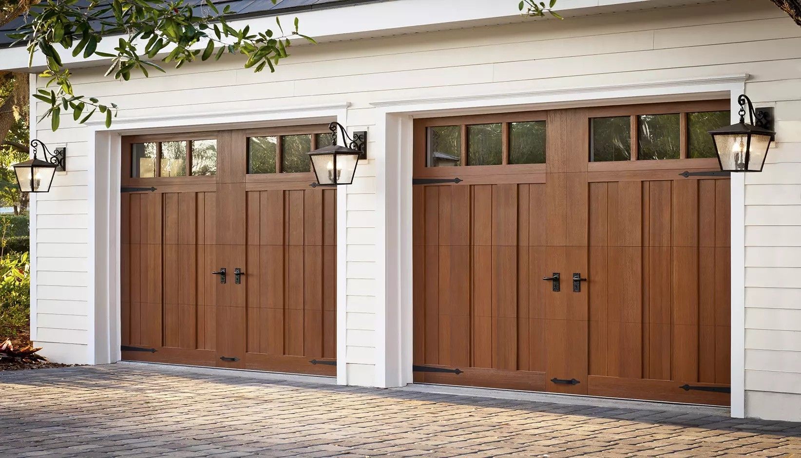Two brown, wooden carriage-style garage doors with windows and black hardware on a house with white horizontal siding.