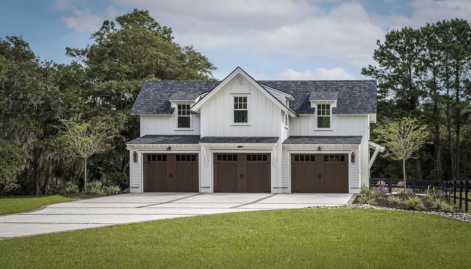A white two-story carriage house with three brown garage doors and a grey roof, set in a sunny, green landscape.