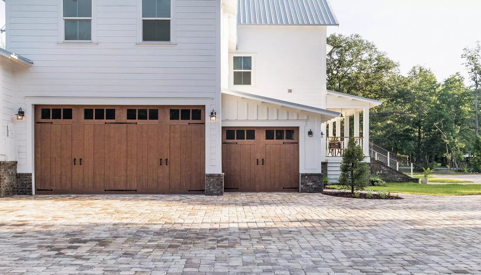 A white farmhouse with two wooden garage doors, a covered porch, and a stone-paved driveway.