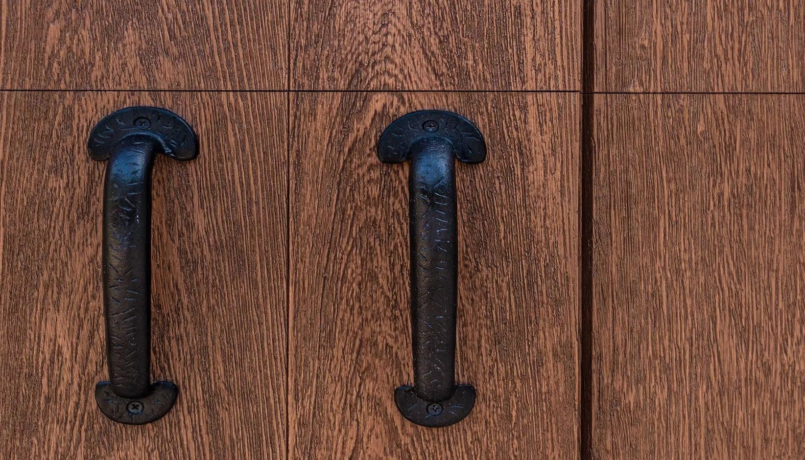 Two dark, rustic metal door handles mounted vertically on a textured, medium-brown wood paneled door.