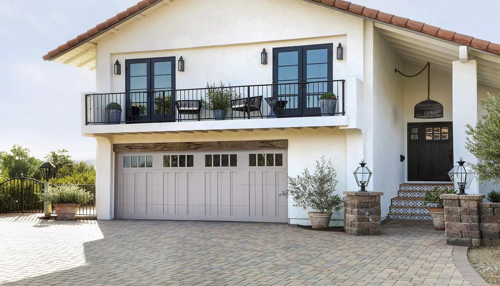 A two-story white house with a beige garage door, upper balcony with black railings, and a cobblestone driveway.