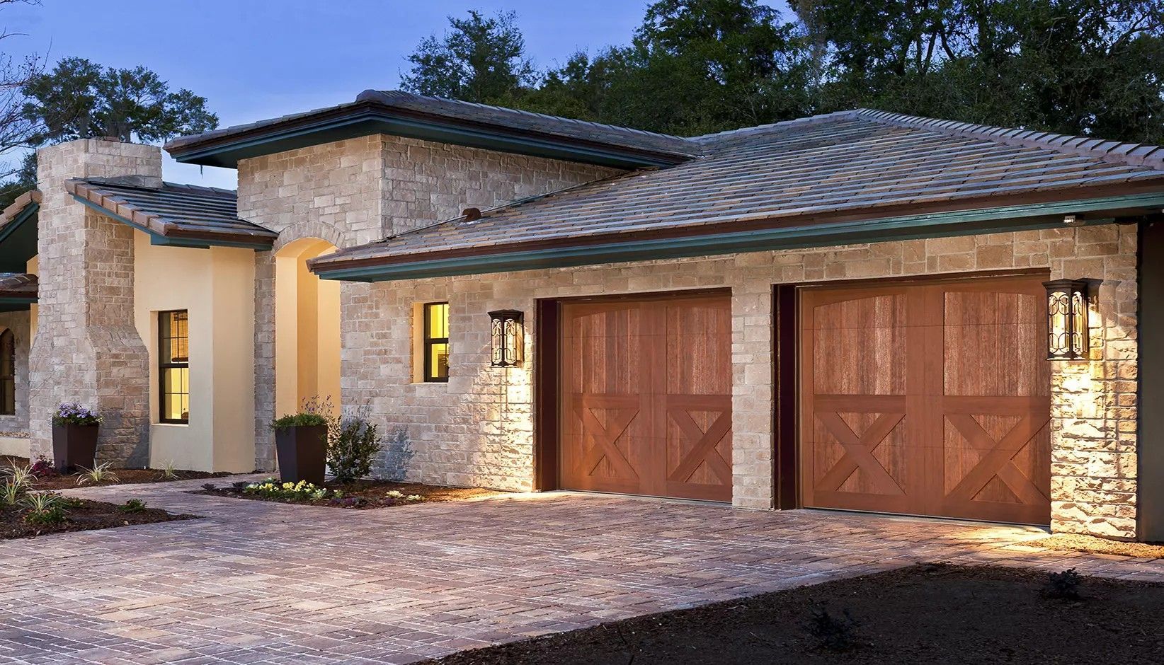 A modern stone house exterior at twilight, featuring a two-car wooden garage and a paved driveway.