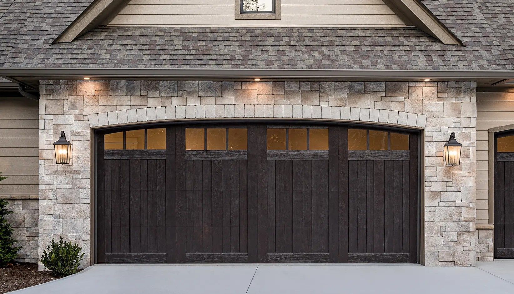 A dark wooden garage door with windows, framed by light-colored natural stone and a shingled roof.
