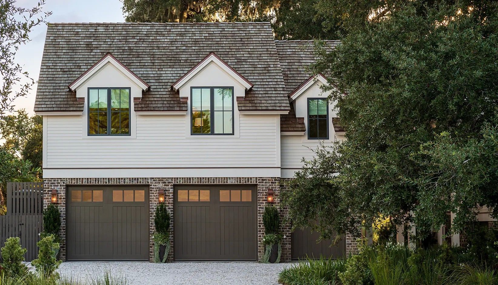 A two-story carriage house with a gray roof, white horizontal siding, and three dark garage doors framed by stone.