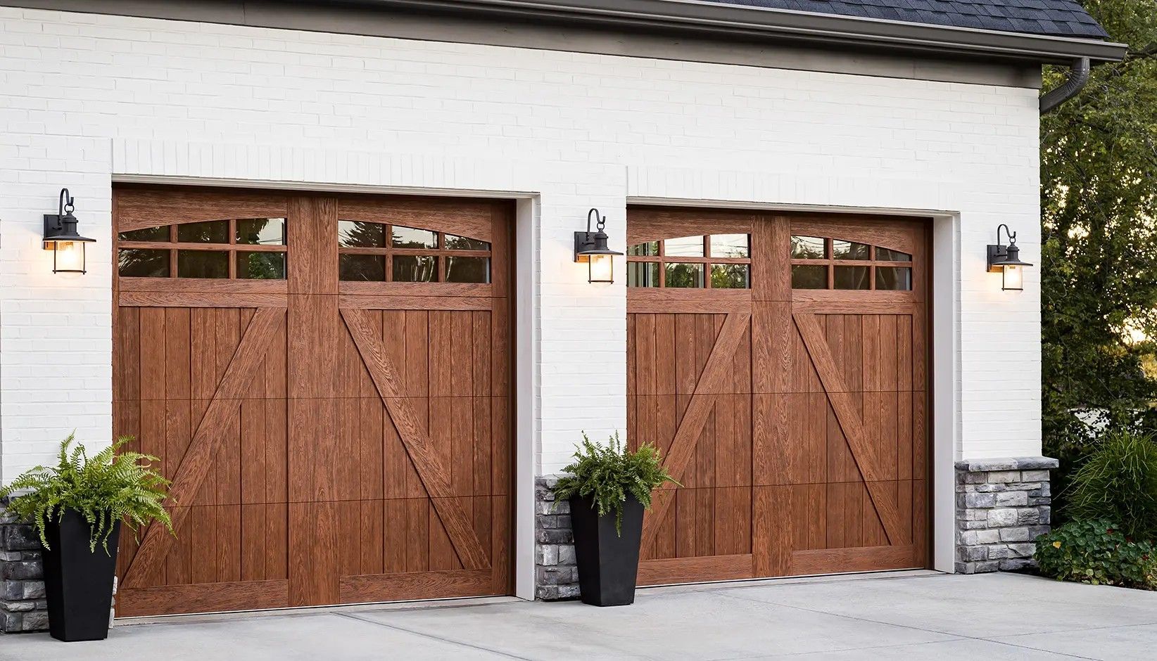 Two wooden garage doors with glass panels and diagonal trim accents on a white brick house with outdoor lanterns.