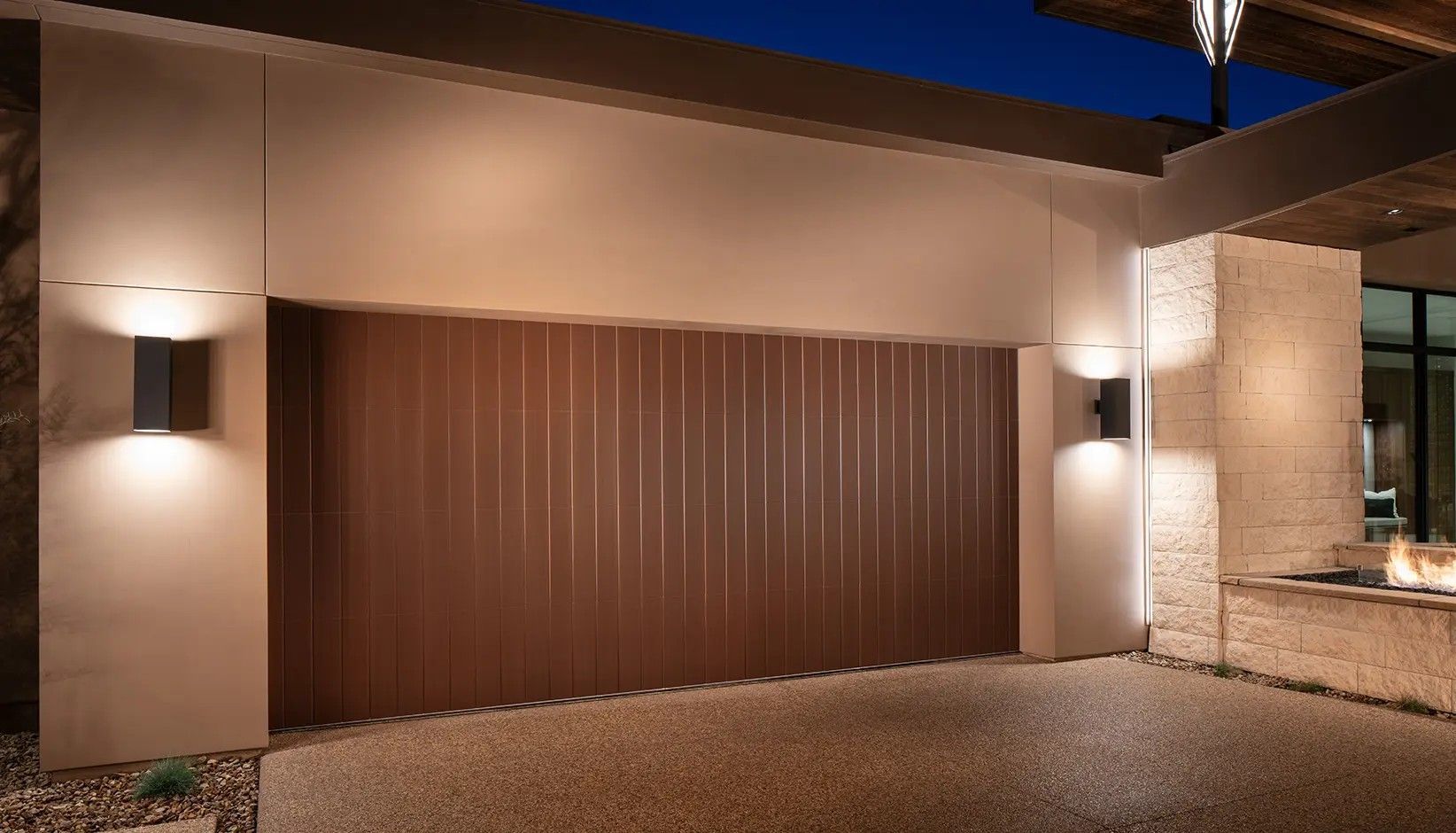 A modern garage door with vertical dark brown paneling, framed by exterior wall sconces at night.
