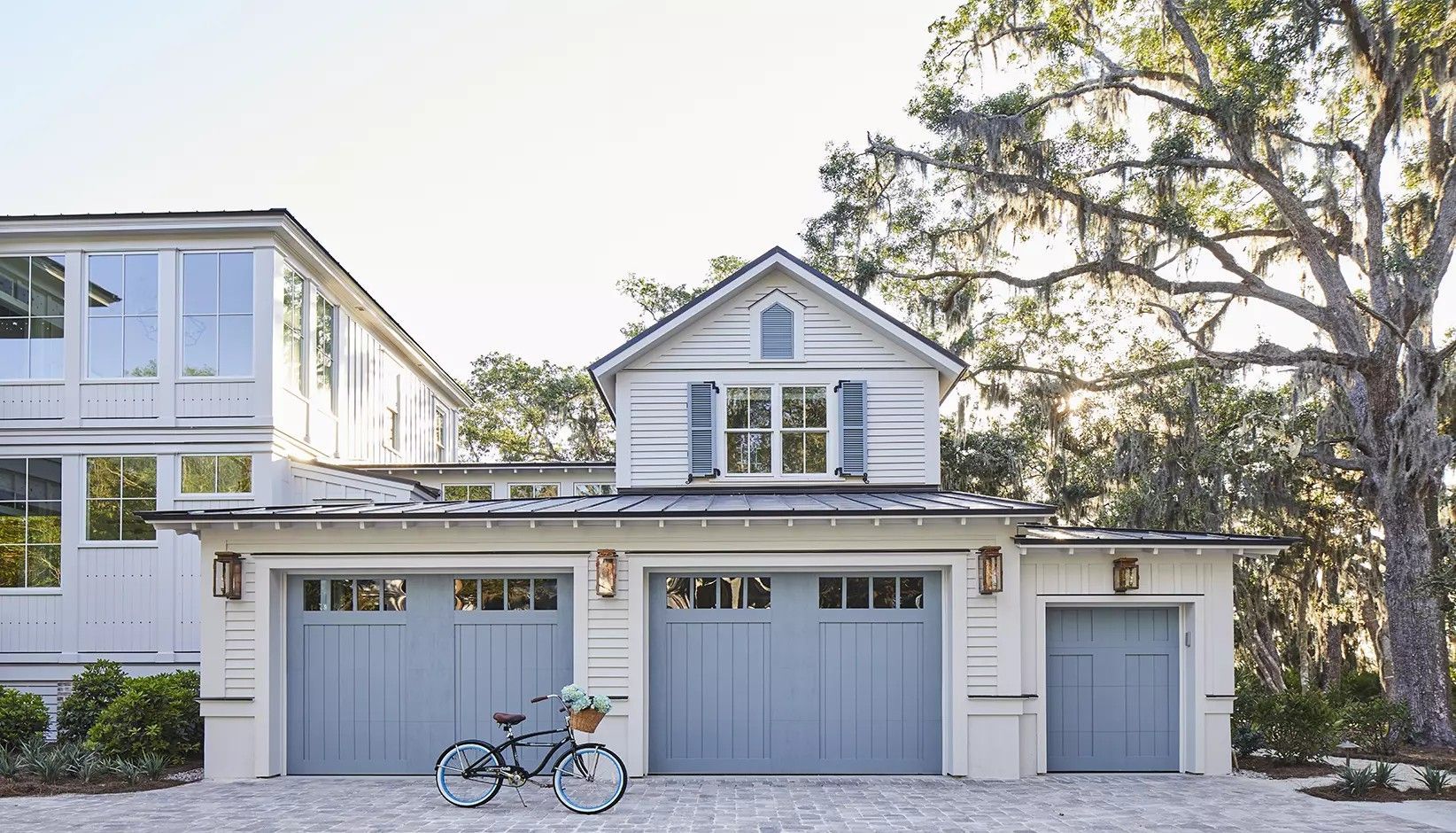 A cream-colored garage with three blue doors and a loft window sits under a large oak tree with a bicycle parked in front.