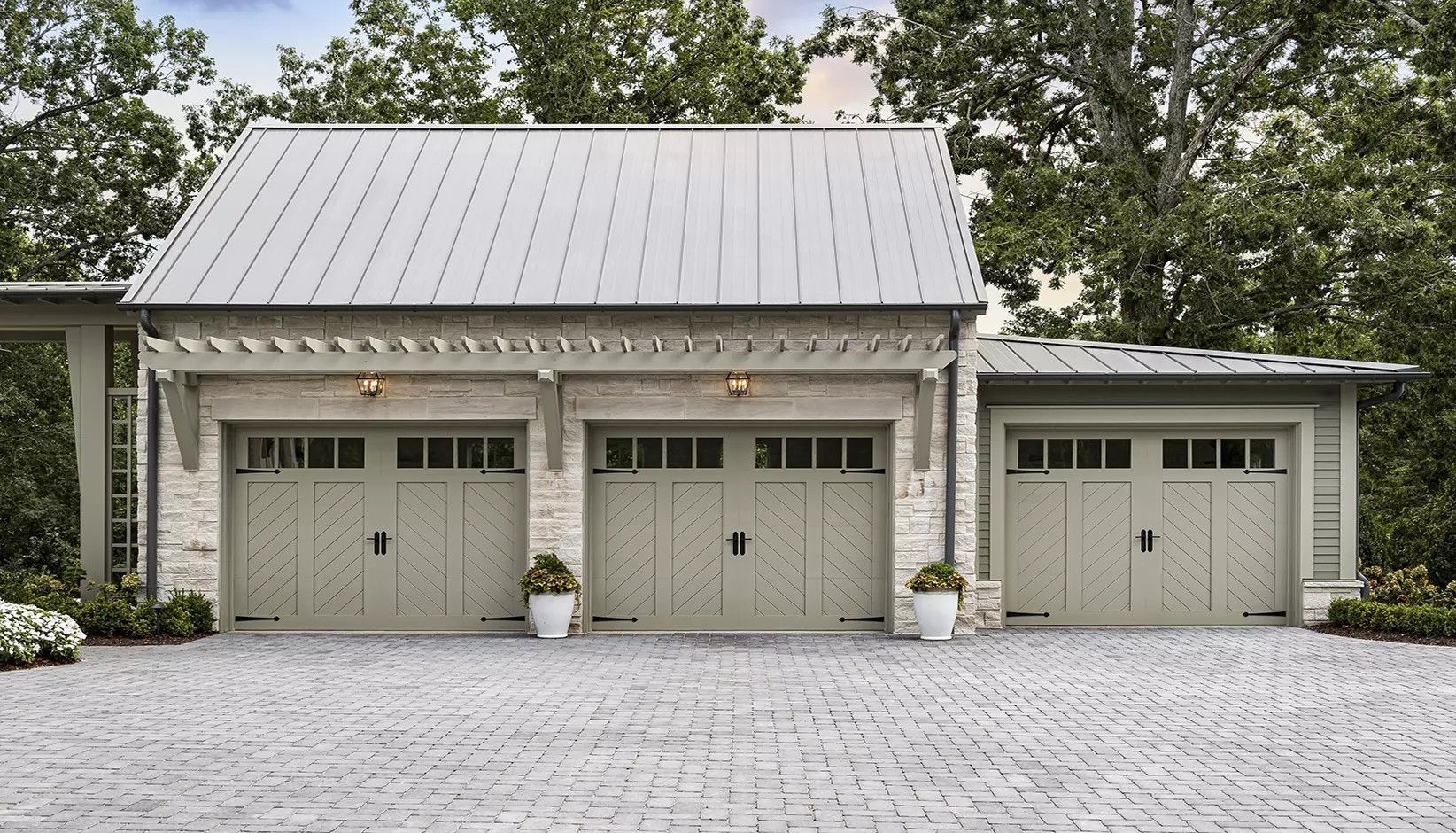 A detached, three-car stone garage with sage green carriage-style doors, a metal roof, and a stone-paved driveway.
