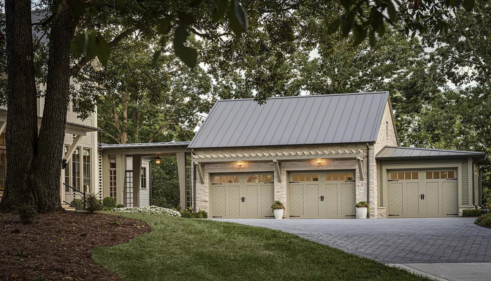 A stone garage with three beige carriage-style doors and a gray metal roof, connected to a home by a covered breezeway.