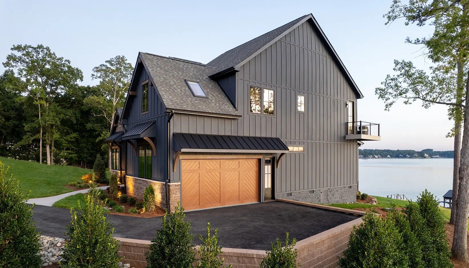 A modern two-story home with dark gray vertical siding, a wooden garage door, and stone accents overlooking a lake.