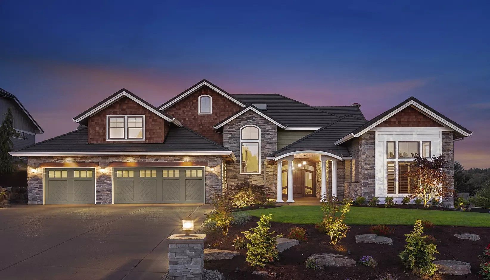 A two-story stone and siding suburban house with a three-car garage and landscaped yard at dusk.