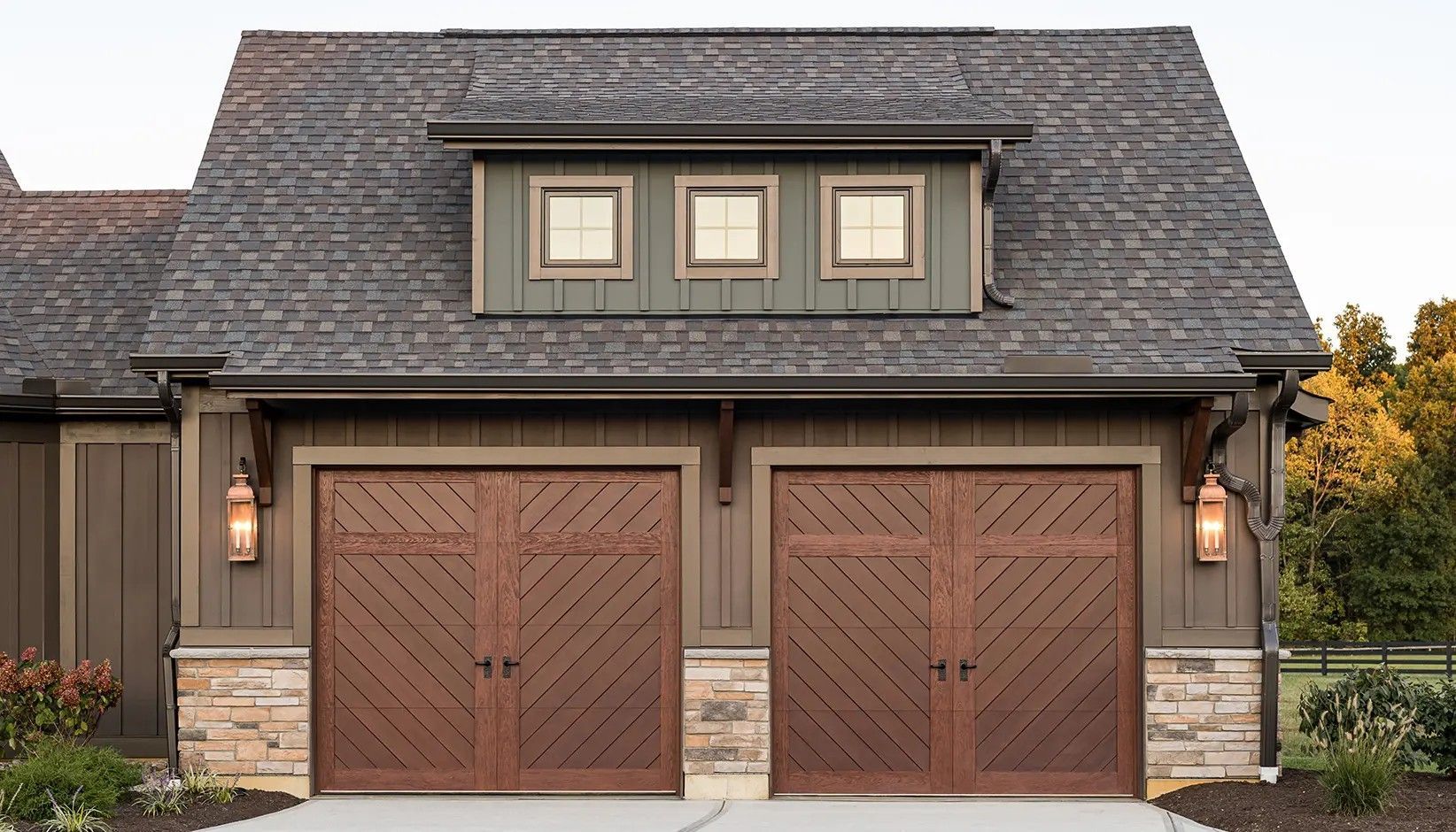 Two wooden carriage-style garage doors with diagonal paneling, stone base siding, and a triple-window dormer above.