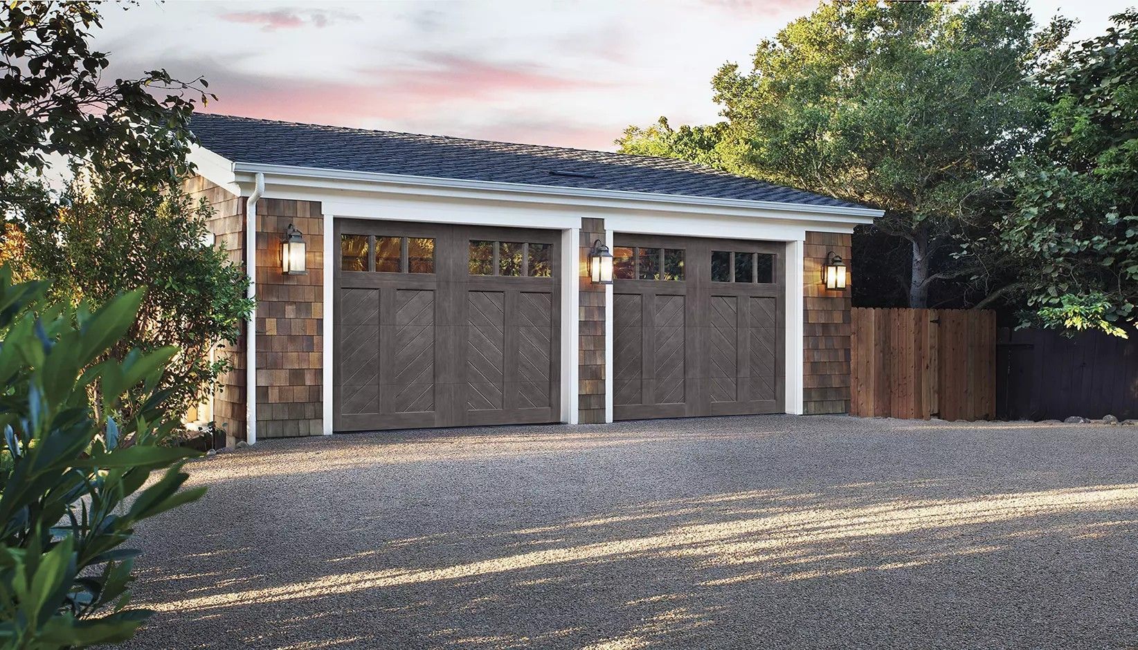 A detached, two-car garage with dark brown wood-paneled doors, stone siding, and gravel driveway at dusk.