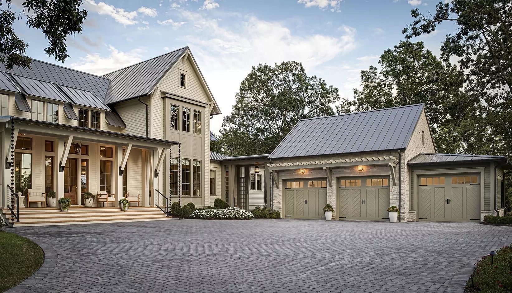 A large, cream-colored house with a metal roof and a matching three-car garage, connected by a stone-paved driveway.