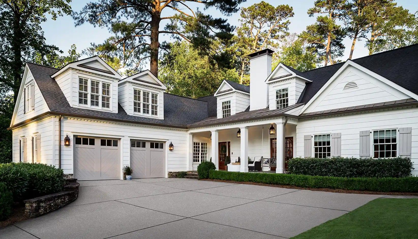 A white, two-story farmhouse-style home with a two-car garage, large porch, and manicured landscaping in a wooded setting.