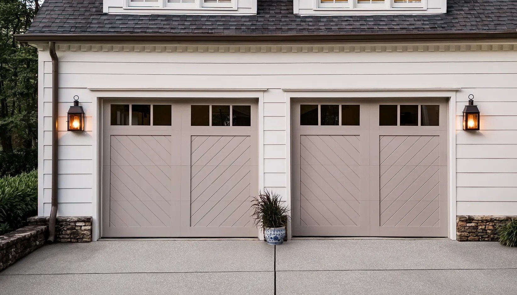Two beige carriage-style garage doors with windows and diagonal wood trim on a white house exterior with outdoor lanterns.