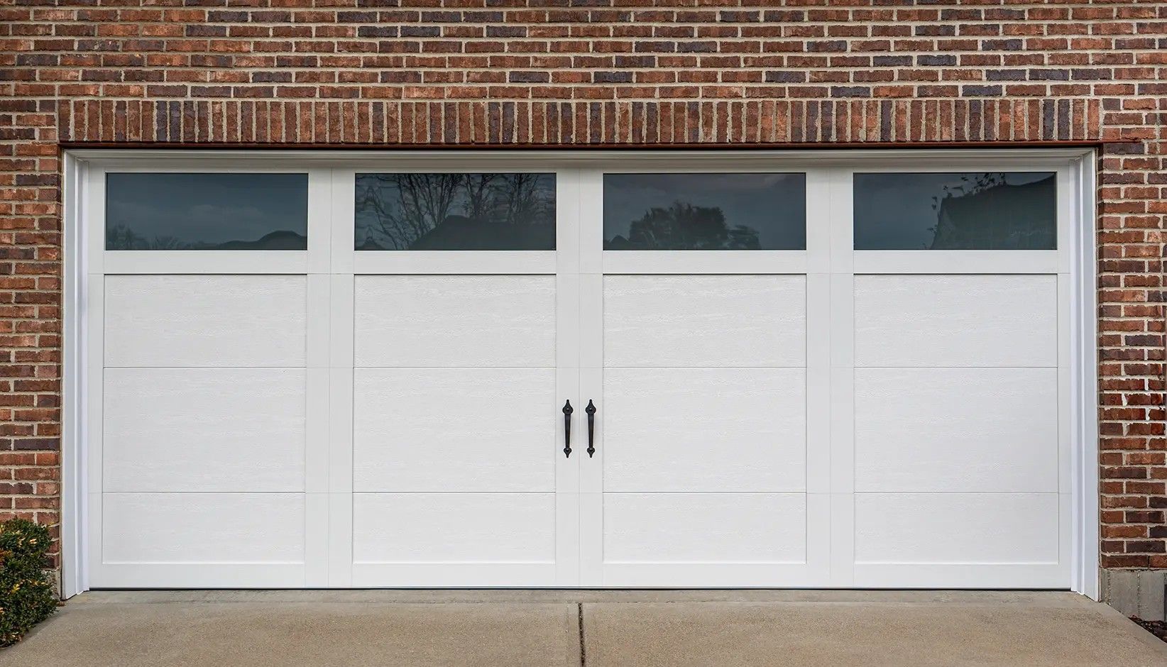 A white, four-panel garage door with top row glass windows and black iron handles installed in a brick exterior wall.