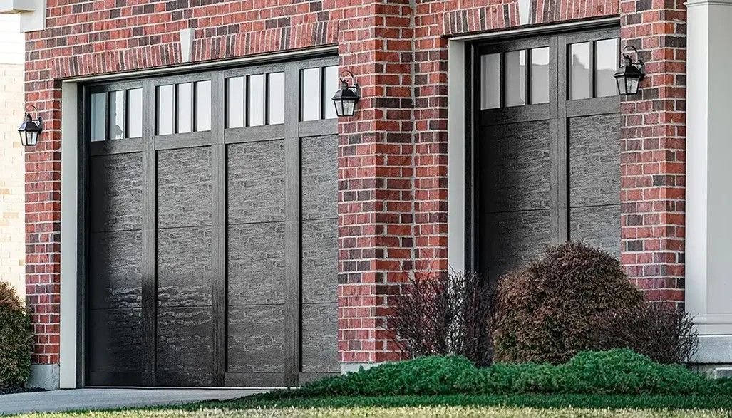 Two dark brown garage doors with rectangular windows set in a red brick wall.