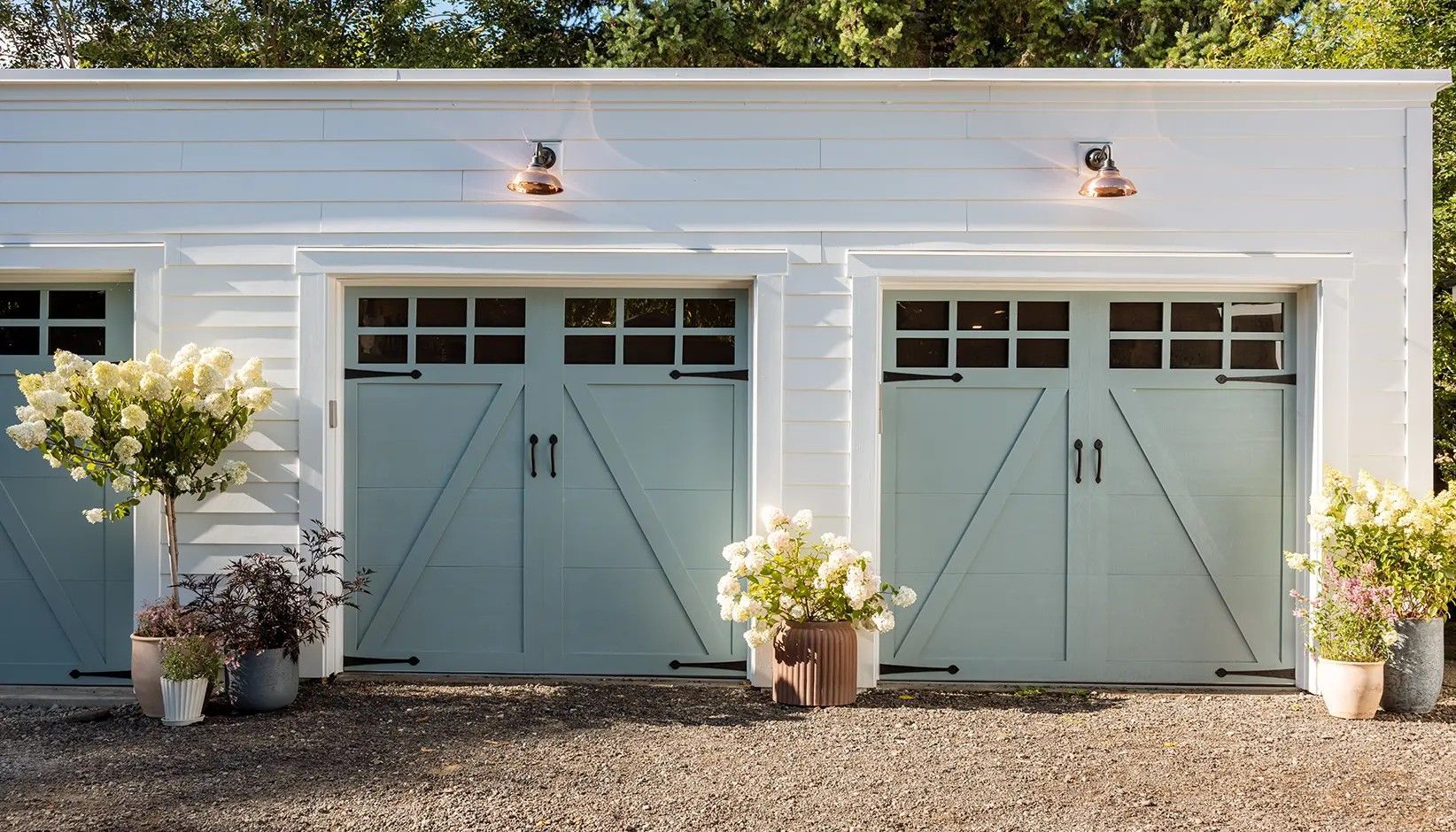 A white garage building with two pale blue carriage-style doors, brass lamps, and potted plants on a gravel driveway.