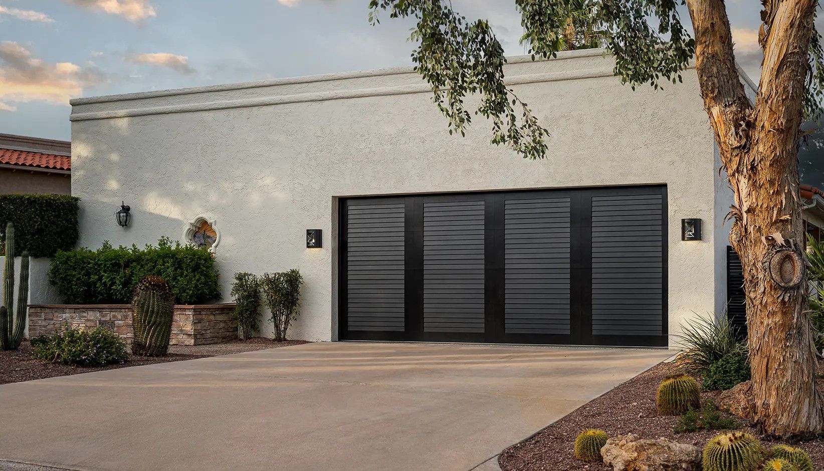 A modern, dark-paneled garage door installed on a textured, light-colored home exterior during sunset.