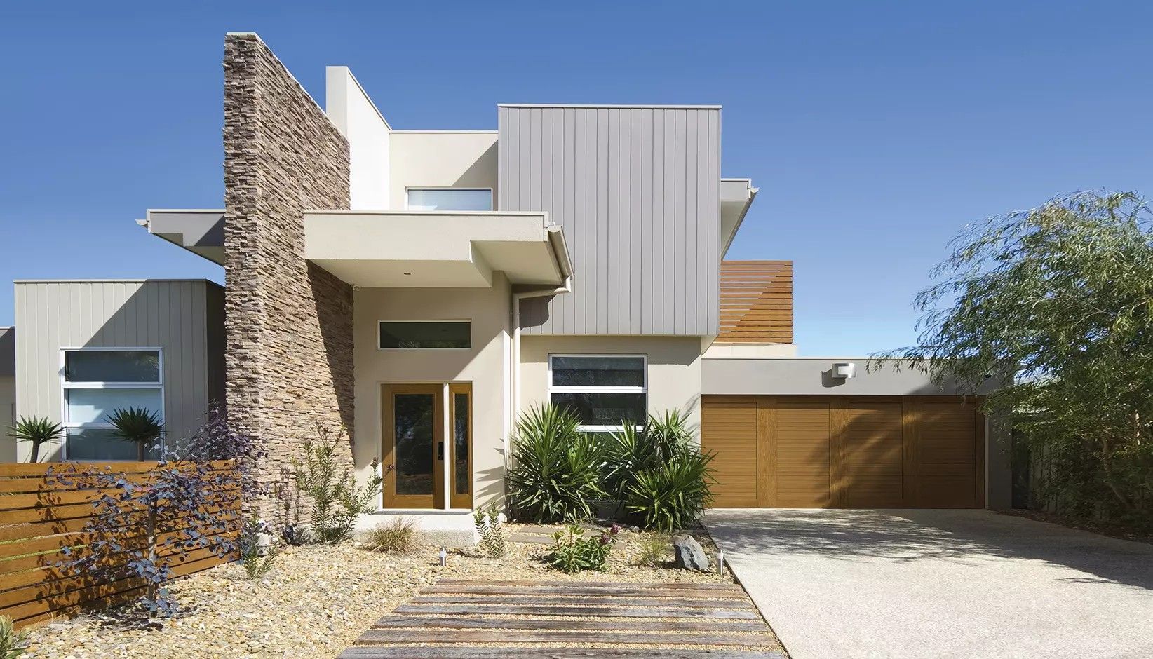 A modern, two-story house with a stone accent wall, wood-paneled garage, and a light-colored exterior under a clear sky.