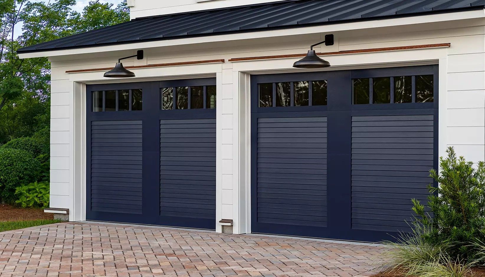 Two navy blue, multi-panel carriage-style garage doors with windows on a white house exterior with a brick driveway.