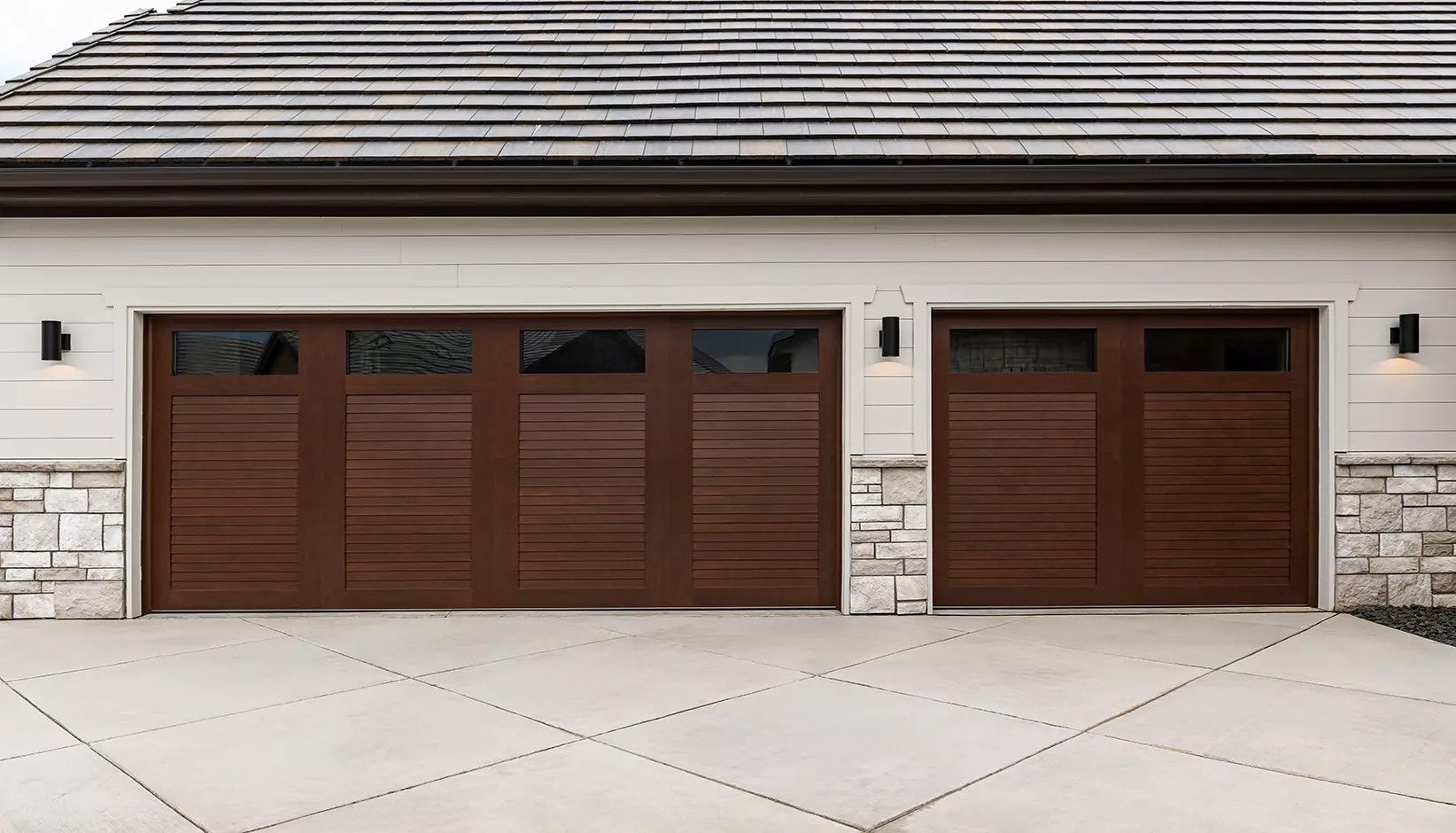 A three-car garage with dark brown horizontal-slatted doors, stone accents, and wall sconces on a white exterior.