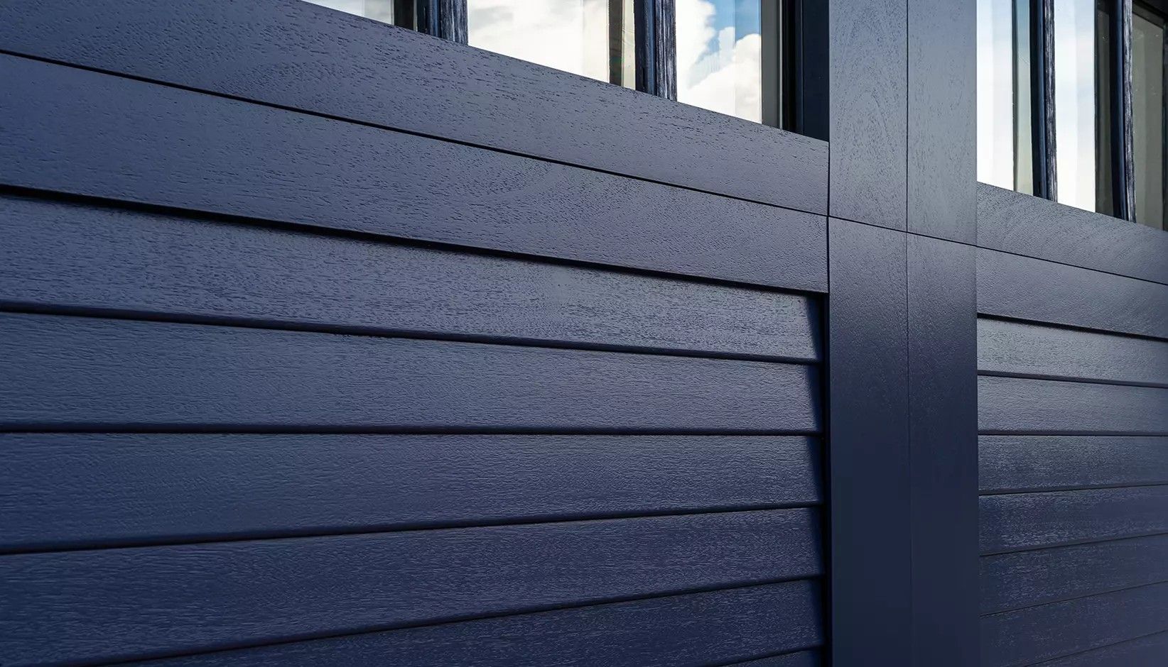 Dark blue horizontal wood-look siding panels meeting at a vertical post, with windows visible in the background.