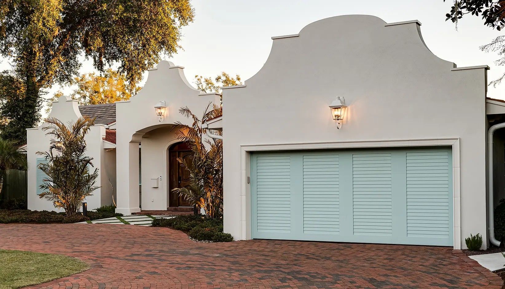 Single-story white home with a mission-style roofline, a light blue garage door, and a brick driveway at twilight.