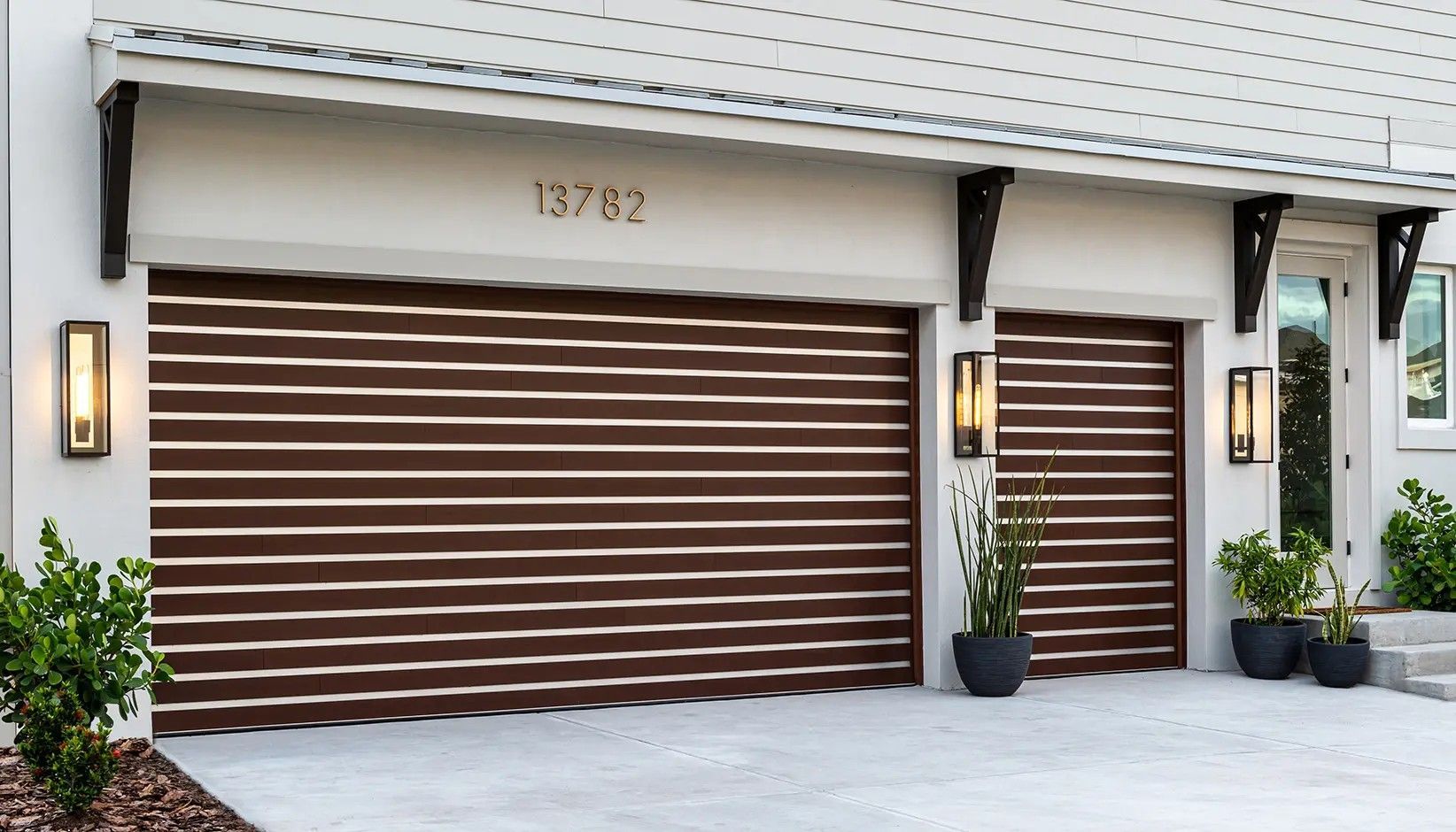 Two brown garage doors with horizontal slats on a modern white house with outdoor lighting and potted plants.