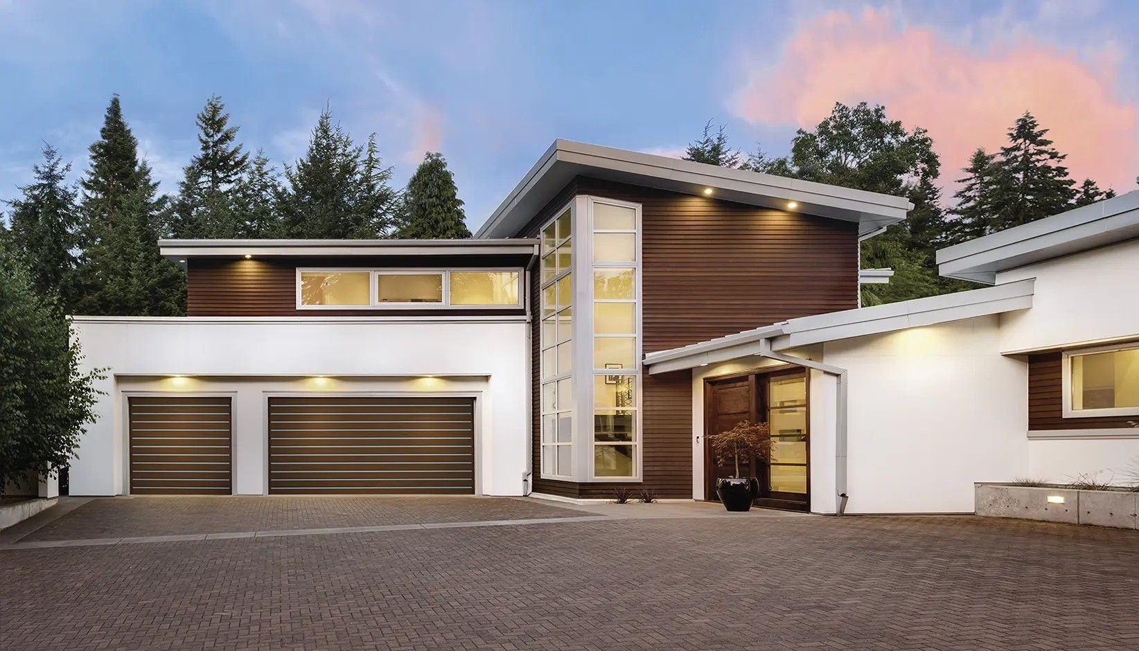 A modern two-story home with a brown and white exterior, a two-car garage, and a tall central glass entry at dusk.