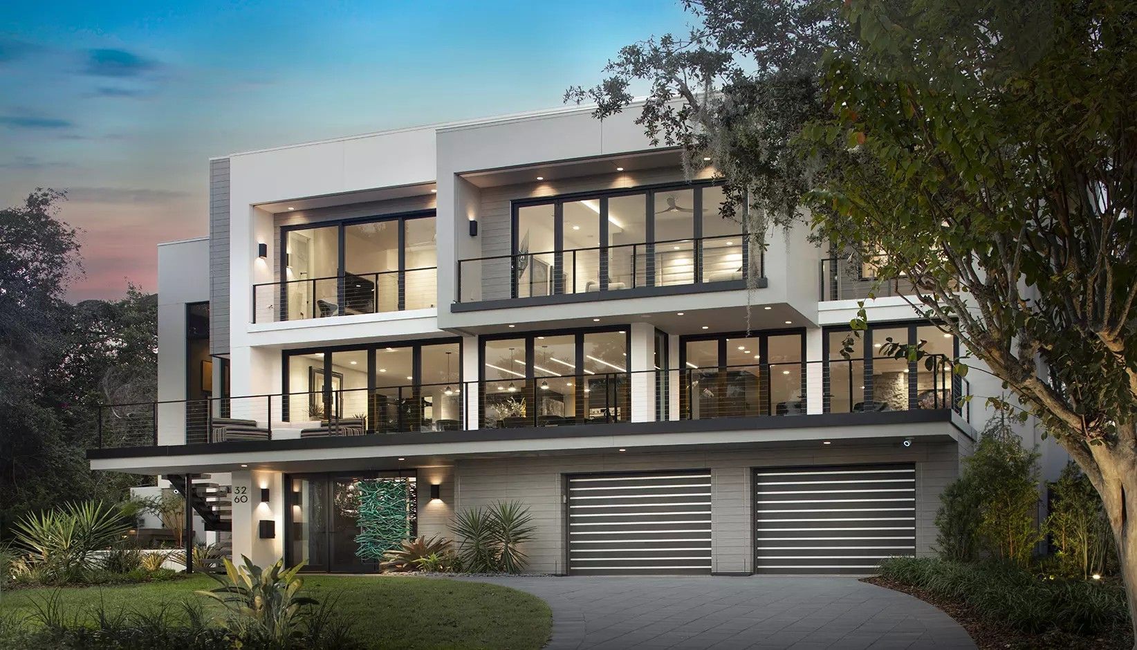 A modern, three-story white home with floor-to-ceiling windows, balconies, and a two-car garage at dusk.