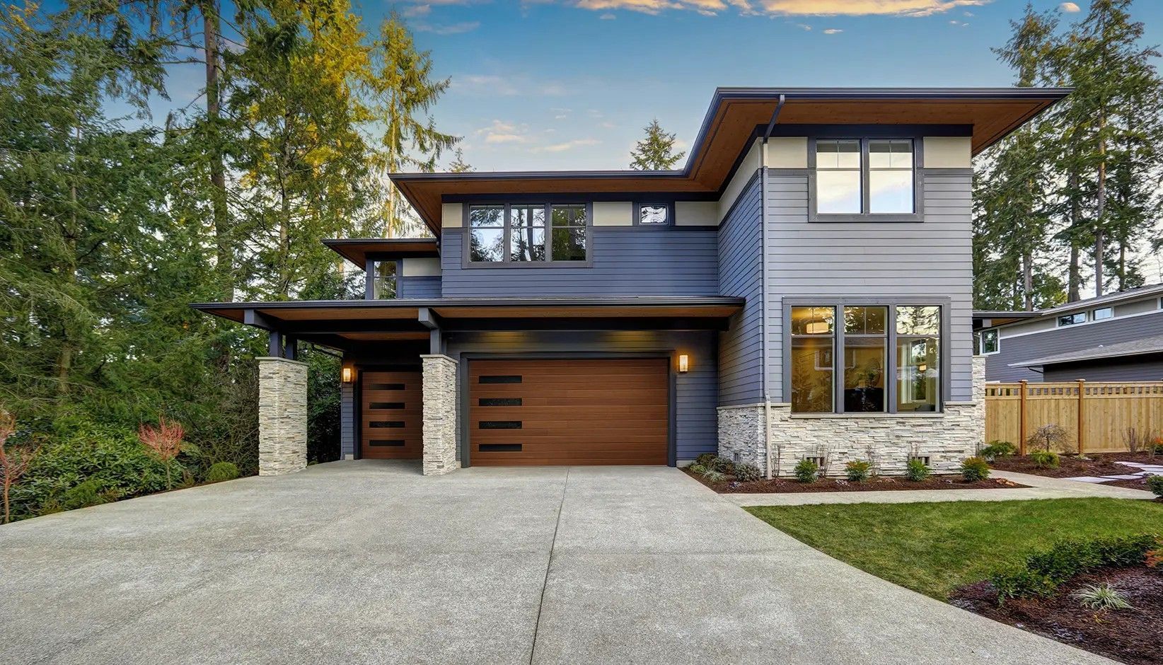 A two-story modern house with a mix of blue siding, wood-paneled garage, stone accents, and a wide concrete driveway.