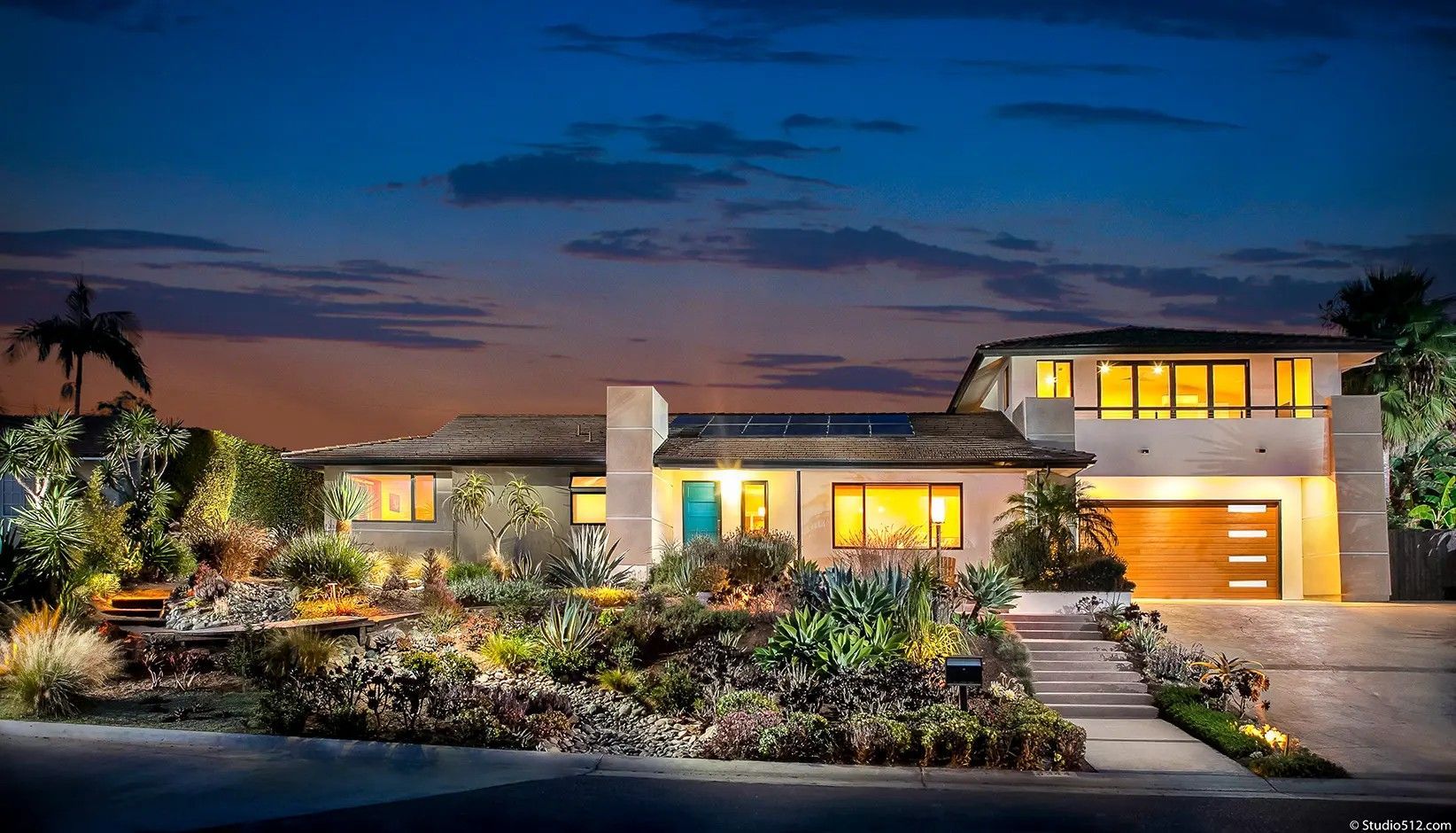 Modern two-story house at dusk with warm interior lighting, a teal front door, and a landscaped succulent front garden.
