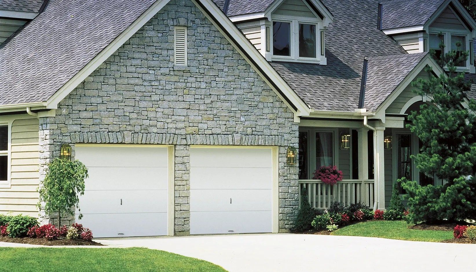 A stone-faced suburban house featuring a two-car garage, gray roof shingles, and a landscaped front porch.