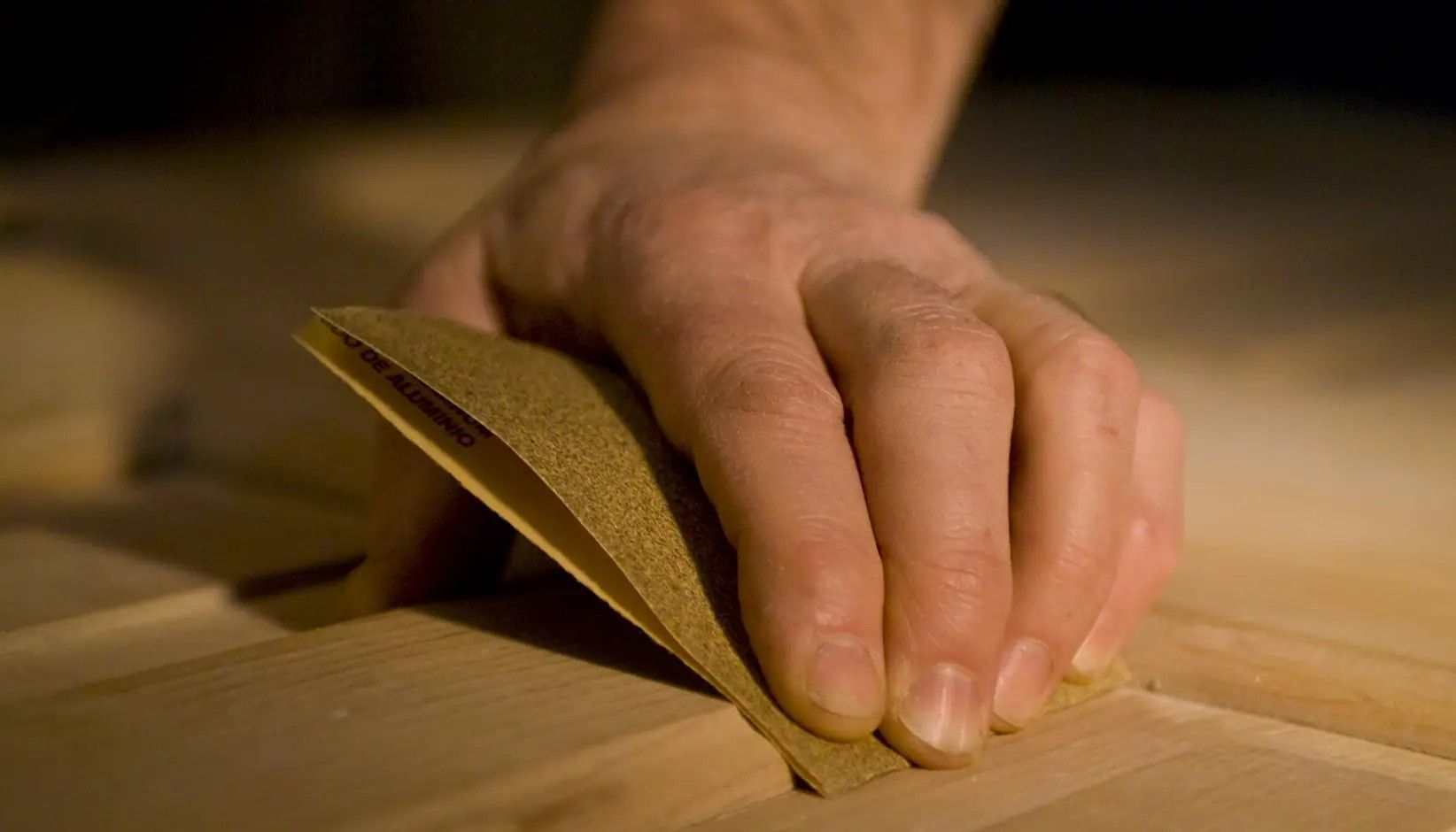 A close-up view of a hand using a piece of tan sandpaper to smooth the surface of a light-colored wooden board.