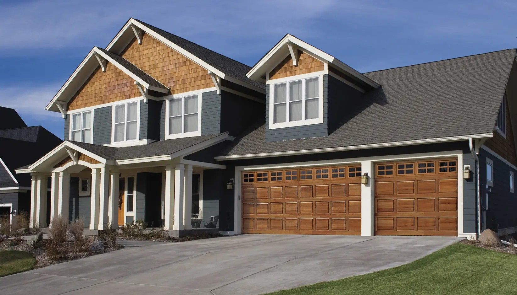 A large, two-story house with dark grey siding, cedar shingle gables, and a two-car wooden garage under a blue sky.