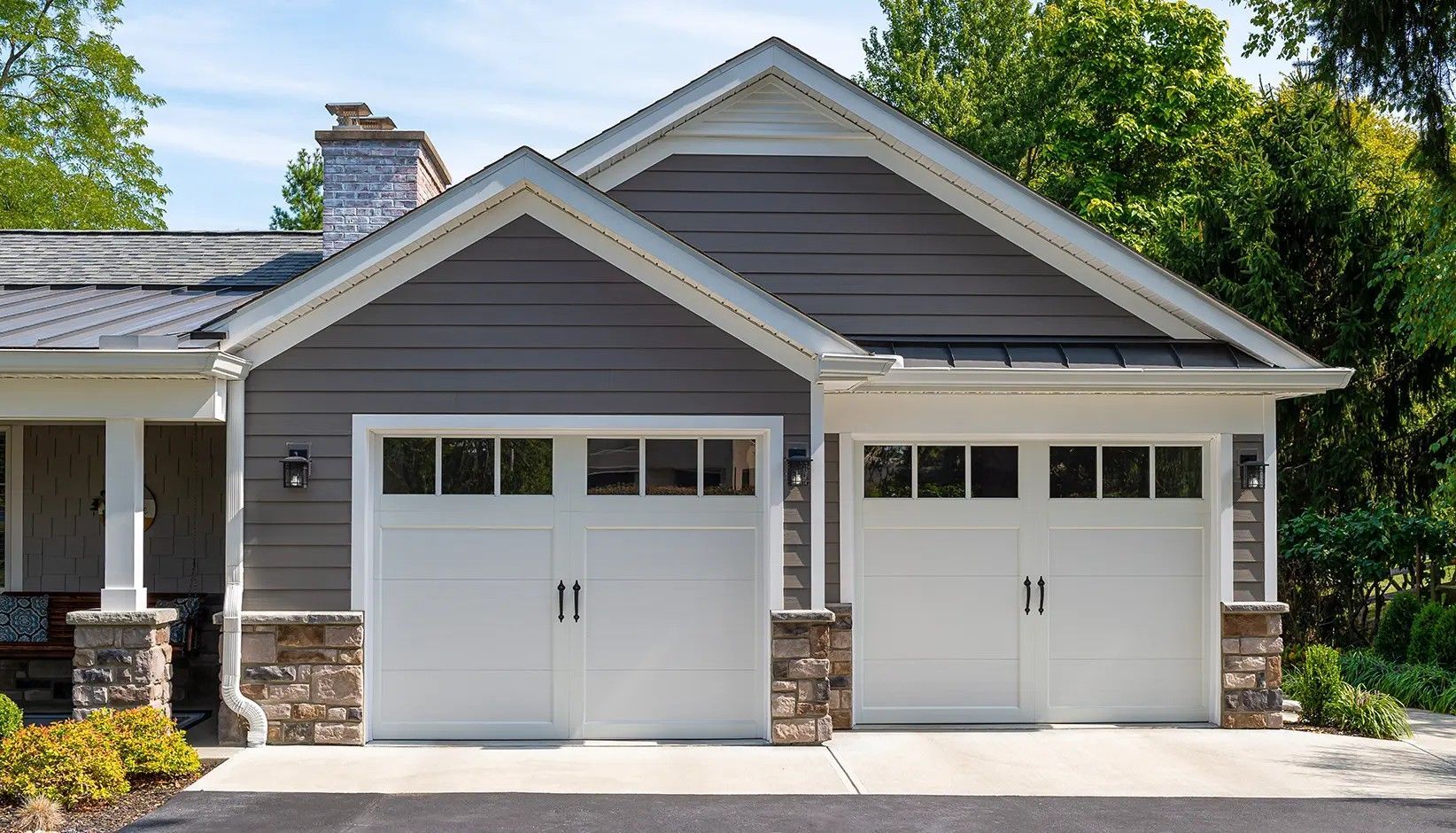 A gray-sided house with two white garage doors, a stone base, and a metal roof surrounded by trees.