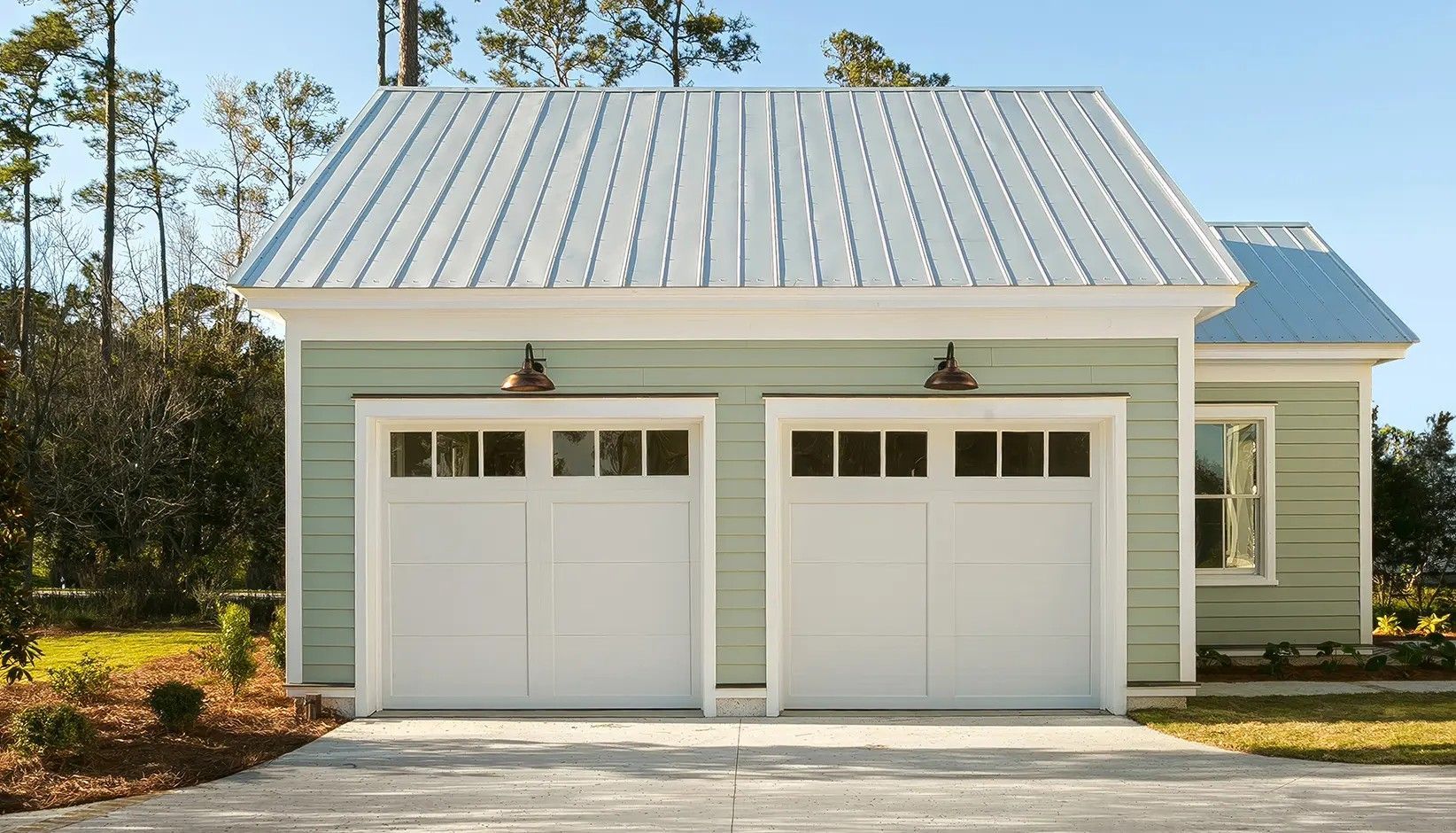 A light green detached two-car garage with a metal roof and white carriage-style doors sits under a clear blue sky.