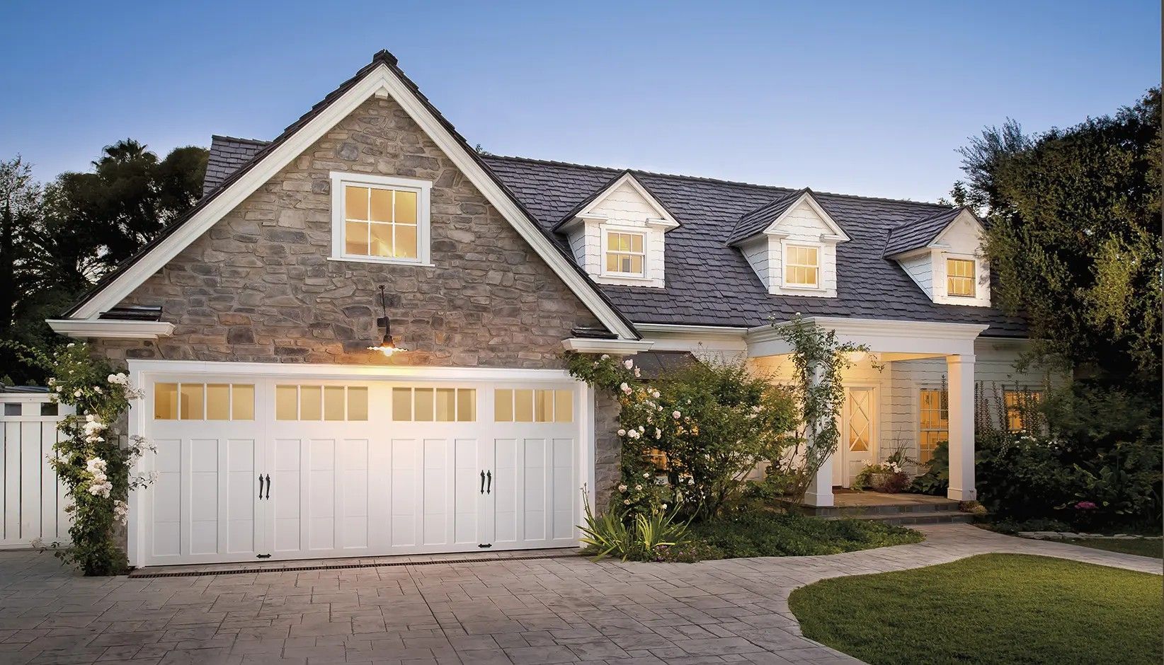 A stone-faced house with a large white three-car garage and dormer windows under a twilight sky.