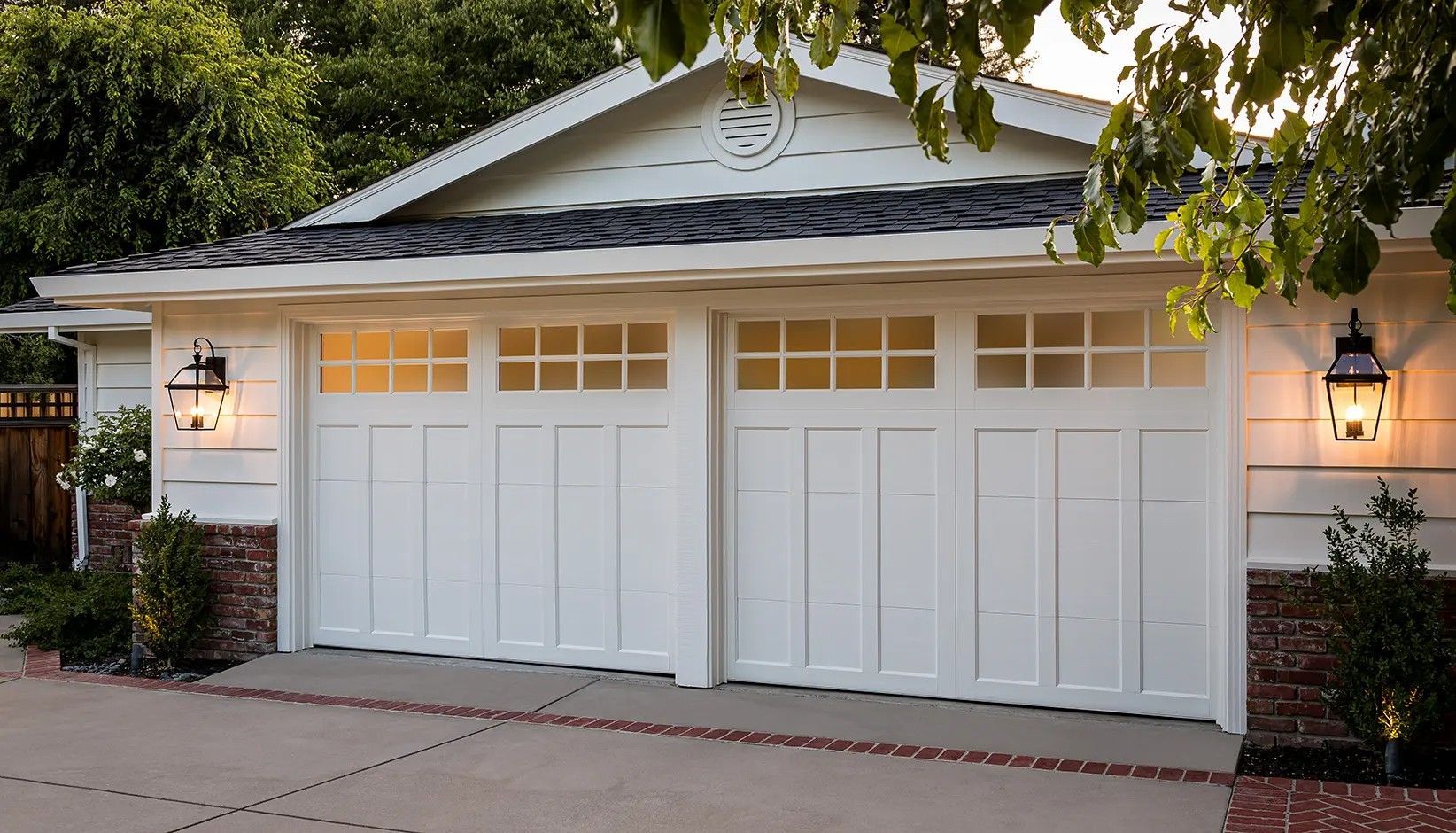 A two-car garage with white carriage-style doors featuring windows, illuminated by wall lanterns on both sides.